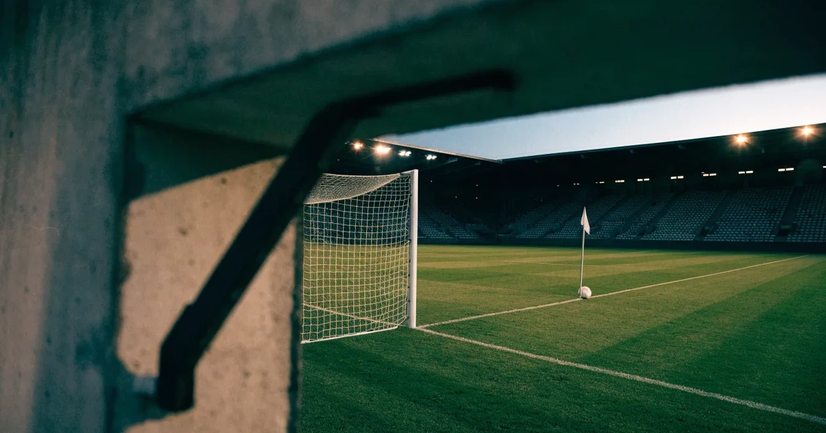 Blick durch ein Stadiontor auf einen grünen Rasen im Abendlicht, ein Ball liegt an der Eckfahne, die Ränge sind leer.