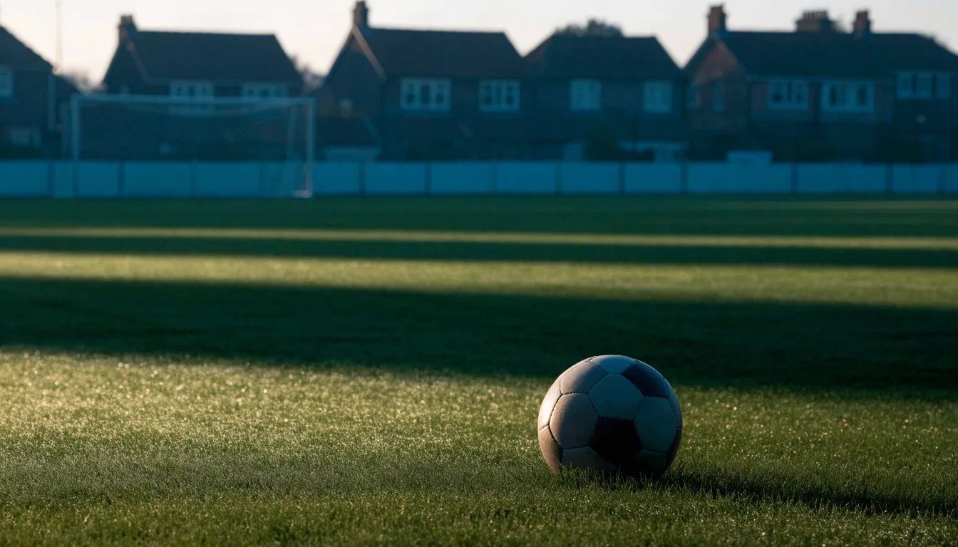 Ein Fußball auf dem Rasen eines leeren Sportplatzes am Rand einer Kleinstadt im Morgenlicht.