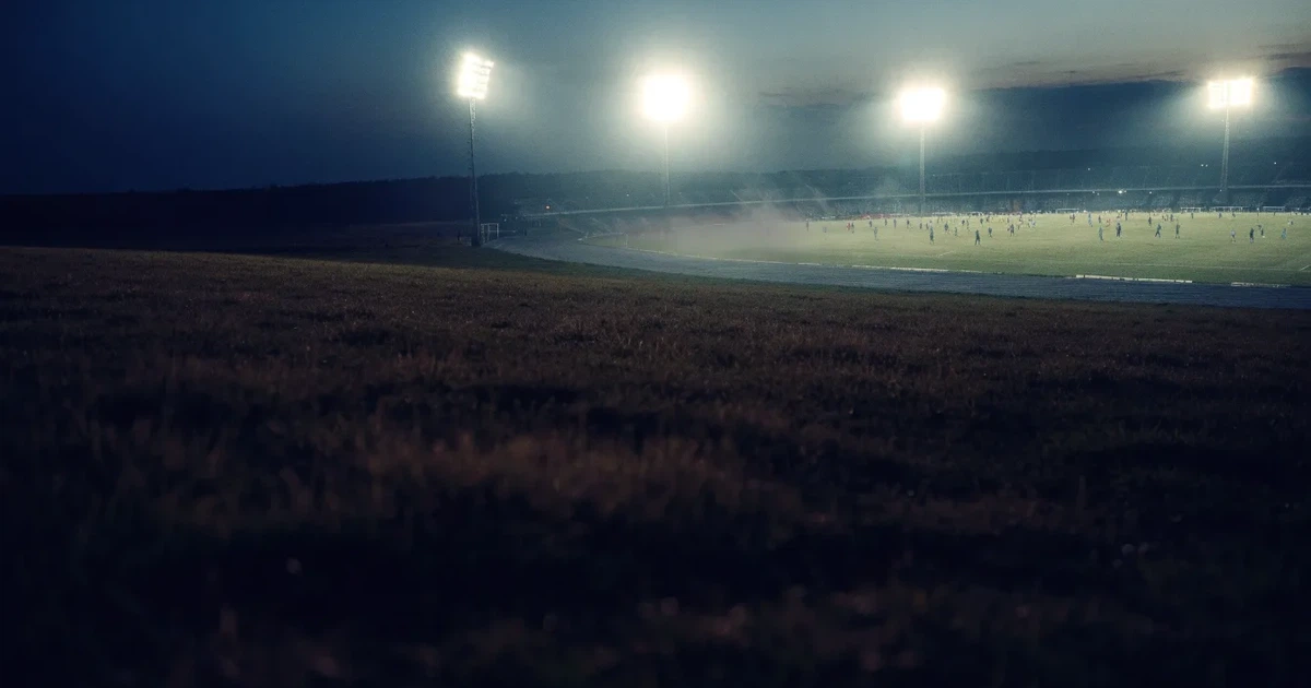 Ein Stadion unter Flutlicht, aus großer Entfernung über eine dunkle Wiese fotografiert, am Himmel zieht Nebel auf.