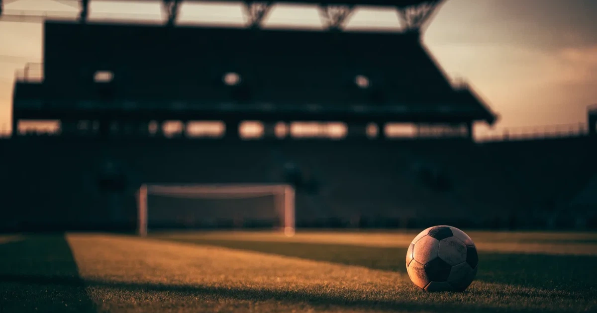 Ein Fußballplatz mit frisch gemähtem Rasen im Abendlicht, ein Ball liegt im Strafraum, im Hintergrund unscharf eine leere Tribüne.