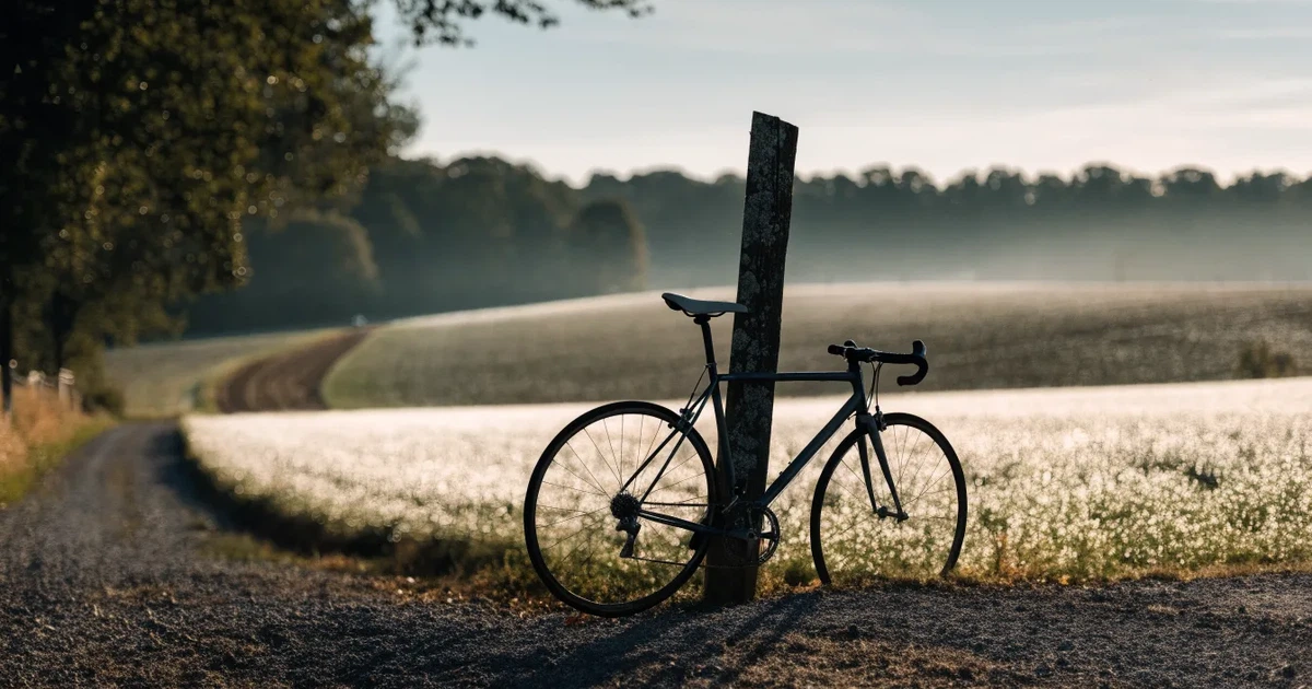 Eine Landstraße zwischen Feldern im Morgenlicht, am Straßenrand lehnt ein Rennrad ohne Aufschrift an einem Pfosten.