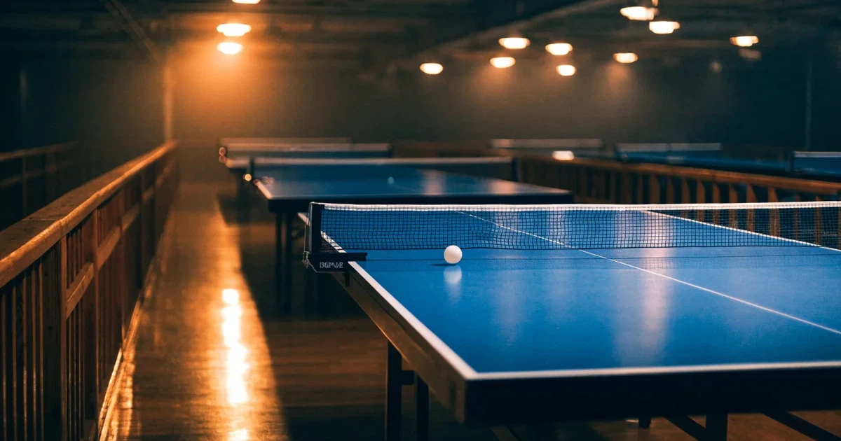 Eine Tischtennishalle aus erhöhter Perspektive mit mehreren blauen Tischen in Reihen unter gedämpftem Hallenlicht, ohne Personen.