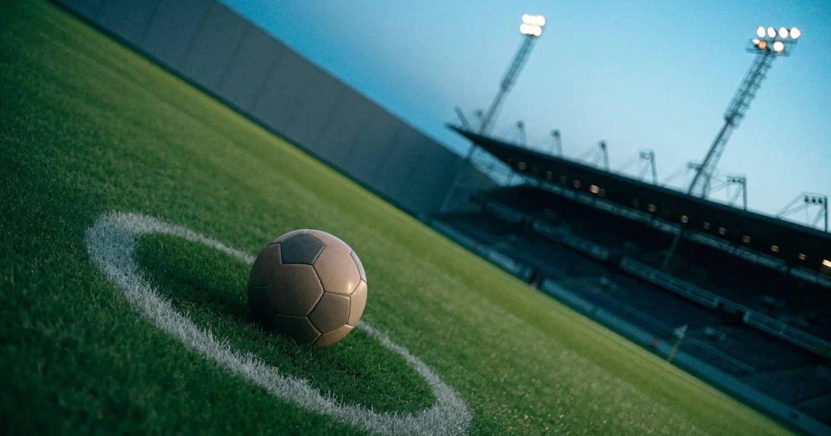 Ein Fußball liegt auf dem Anstoßpunkt eines leeren Rasenplatzes, im Hintergrund eine Tribüne und Flutlichtmasten in der Abenddämmerung.