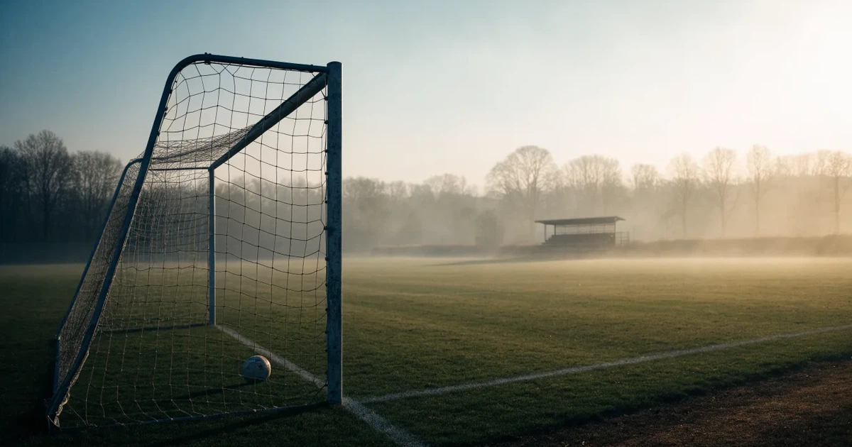 Ein Fußballtor mit Netz steht im Morgennebel auf einem leeren Rasenplatz, im Hintergrund die Silhouette einer kleinen Tribüne.