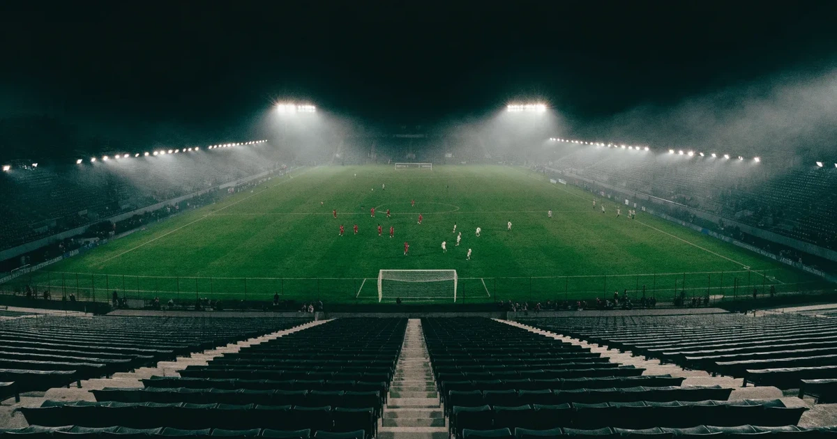 Ein leeres Stadion unter Flutlicht aus großer Entfernung gesehen, Dunst liegt über dem Rasen, die Tribünen sind nur als Lichtpunkte erkennbar.
