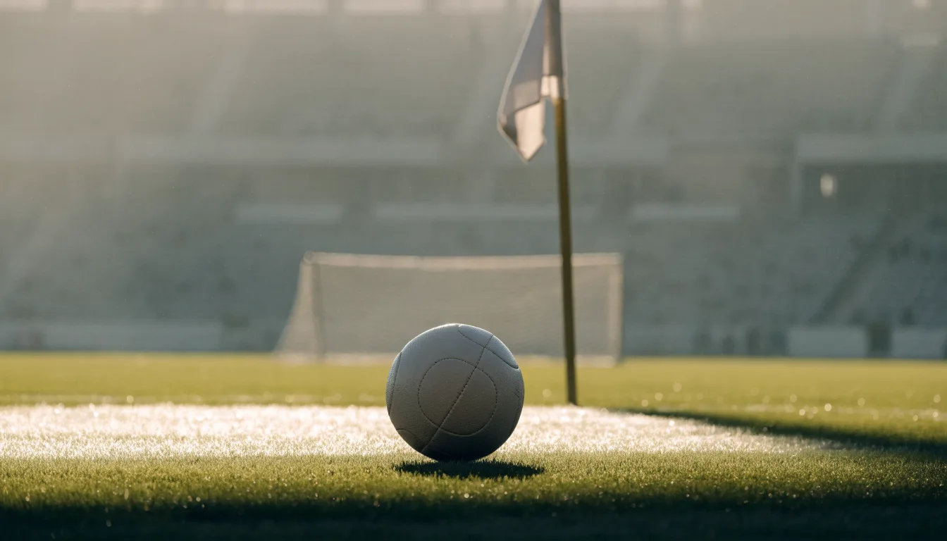 Ein Fußball liegt auf Rasen neben einer Eckfahne ohne Aufdruck auf einem leeren Platz im Morgenlicht.