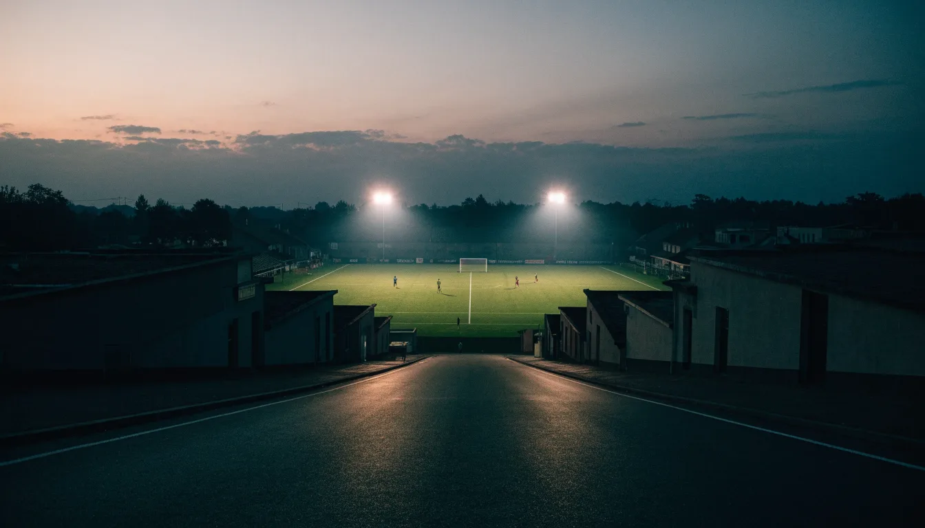 Ein Stadion unter Flutlicht aus großer Entfernung, davor eine leere Straße in der Abenddämmerung.