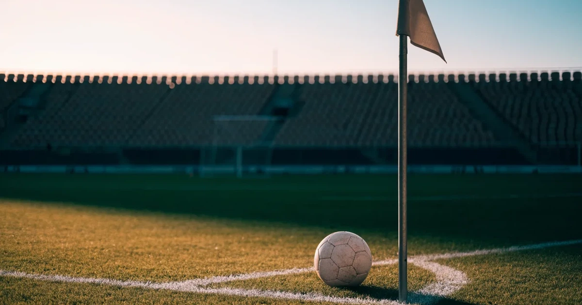 Ein Fußball liegt am Viertelkreis an der Eckfahne eines Rasenplatzes, im Hintergrund leere Tribünen in warmem Abendlicht.